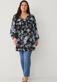 Zwarte bloemetjesblouse met lange mouwen, voorzien van blauwe en groene bloemen, een V-hals en geplooide details bij de buste. Gecombineerd met blauwe jeans.