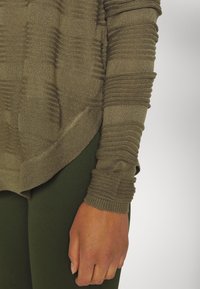 Olivgrüner, gestrickter Pullover mit strukturierten Streifen an den Ärmeln, kombiniert mit dunkelgrünen Leggings. Verfügt über eine lockere Passform und gerippte Kanten.