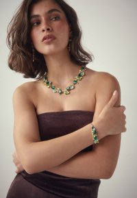 Emerald green and gold jewelry set featuring a beaded necklace and bracelet with various gem shapes and textures, worn with a strapless top.