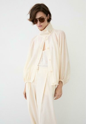 Frau trägt eine cremefarbene, hochgeschlossene Bluse mit Knöpfen und Baumschulärmeln, passende hoch taillierte Hose, braune Sonnenbrille und eine Halskette.