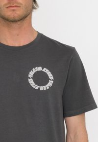 Camiseta de algodón gris oscuro con mangas cortas, presenta un gráfico blanco circular en el pecho, cuello redondo y una textura suave.
