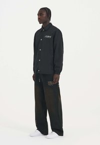 Chaqueta negra de manga larga con botones a presión y detalles de logo; combinada con pantalones vaqueros oscuros de corte holgado que presentan texto bordado y acentos.
