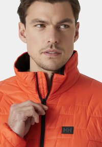 Chaqueta acolchada de color naranja brillante con cremallera negra y forro interno. Presenta un cuello alto y un pequeño logo negro en el pecho.