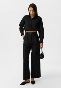 Zwarte cropped button-up blouse met lange mouwen, gecombineerd met hoge zwarte wijdpijp broeken. Het model houdt een kleine zwarte handtas vast.