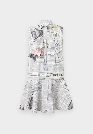 Μαύρη γυναίκα που φοράει αμάνικο φόρεμα με τύπωμα εφημερίδας και διακοσμητικά βολάν, στέκεται με το ένα χέρι χαλαρό, φοράει μαύρα ψηλοτάκουνα σανδάλια.