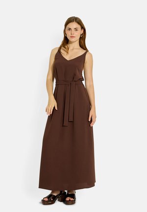 Mujer con vestido marrón largo sin mangas con cinturón en la cintura y sandalias negras de plataforma, de pie frente a un fondo claro liso.