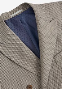 Blazer beige et marron à motif pied-de-poule avec larges revers, bouton marron et doublure intérieure bleu foncé à motif géométrique.