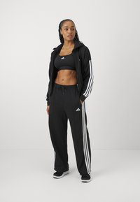 Svart Adidas-träningsoverall med en zip-up jacka med vit tre-stripes design och matchande träningsoverall. Inkluderar en svart sport-bh.