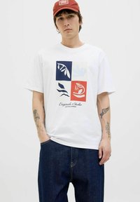 Hvid kortærmet t-shirt med et grafisk design bestående af fire farvede firkanter, der viser bladmønstre i blå, lys blå og rød.