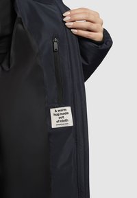 Tengerész dzseki sima külső anyagból, cipzáras zsebbel és egy címkével, amely azt mondja: "Egy meleg ölelés, amit szövetből készítettek." Kontrasztos bélés látható.