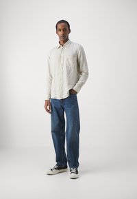 Camisa de lino color beige claro con botones al frente y bolsillo en el pecho, combinada con jeans de mezclilla azules y zapatillas en blanco y negro. Diseño simple, ajuste casual.
