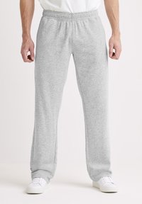 Man draagt lichtgrijze joggingbroek en witte sneakers, staat tegen een effen achtergrond met handen ontspannen langs het lichaam.