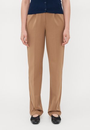 Brune skreddersydde bukser med rett passform, frontfold, og sidelommer, sammen med svarte slip-on sko.