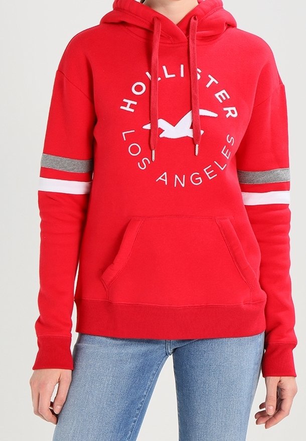 Sweat à capuche rouge avec des rayures blanches et grises sur les manches, texte « Hollister Los Angeles » et logo d’oiseau sur la poitrine, porté avec un jean bleu.