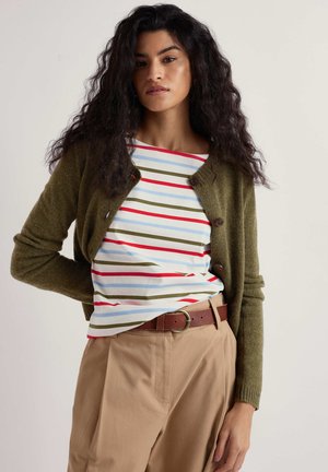 Kobieta z długimi kręconymi czarnymi włosami, ubrana w oliwkowozielony kardigan, koszulę w paski w kolorach czerwonym, niebieskim i zielonym, beżowe spodnie oraz brązowy pasek.