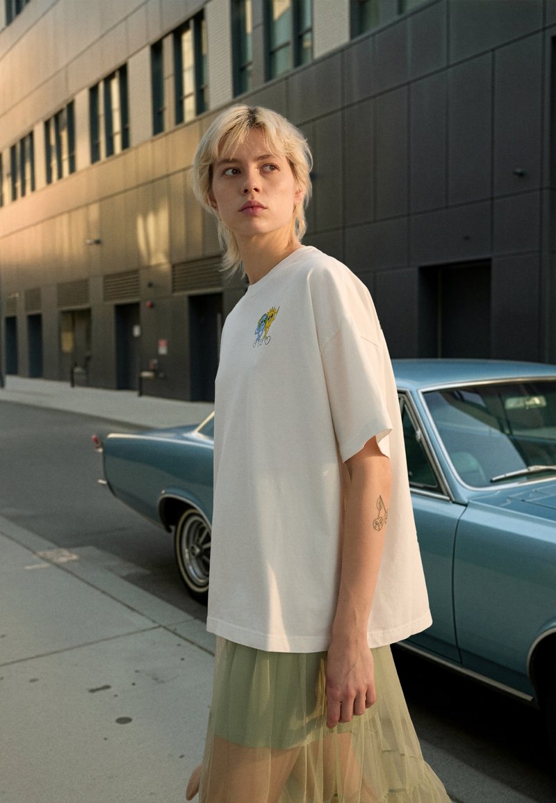 White oversized t-shirt with a graphic print on the left chest and a translucent green midi skirt, set against a vintage blue car.