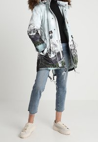 Pitkä sininen parkatakki, jossa vuoristokuvio, turkiksella vuorattu huppu, joustavat kiristysnauhat ja vetoketjulliset taskut, yhdistetty vaaleisiin farkkuihin ja paksupohjaisiin tennareihin.