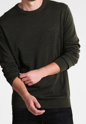 Pull en tricot vert foncé avec des poignets et un ourlet côtelés, arborant un logo discret sur la poitrine. Le matériau a une finition douce et texturée.