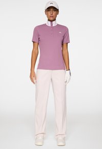 Kurzärmliges, lavendelfarbenes Golf-Polo mit hohem Kragen und Knopfdetail. Kombiniert mit hellrosa Hosen und weißen Sneakers, gehalten mit einem Golfhandschuh.