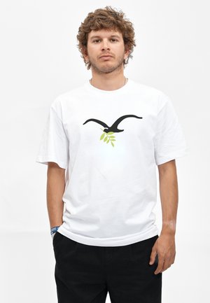 Junger Mann mit lockigem Haar, der ein weißes T-Shirt mit einem schwarzen Vogel trägt, der einen grünen Olivenzweig hält, schwarze Hose und ein blaues Armband.