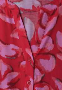 Rød bluse med store abstrakte pink og lilla blomsterprint, med V-hals og to synlige røde knapper foran.