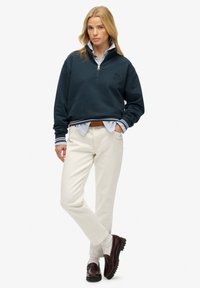 Pull marin avec un col zippé, arborant un logo, superposé sur une chemise rayée bleue et blanche, associé à un pantalon crème et des chaussures bordeaux.