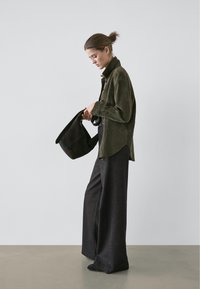 Chemise en daim vert olive, coupe décontractée avec un col montant ; large pantalon gris foncé ; tenant un sac noir ; fond minimaliste.