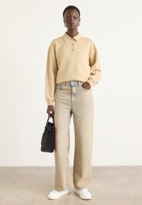 Polo shirt a maniche lunghe beige con colletto, abbinato a jeans a gamba larga in denim chiaro e sneakers bianche. Una borsa nera è tenuta in una mano.