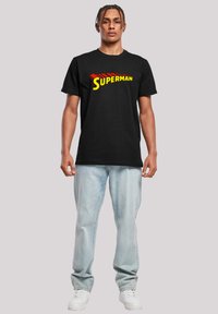 Juodas medvilninis marškinėliai su „SUPERMAN“ tekstu raudona ir geltona spalvomis. Derinami su šviesiai mėlynais džinsais ir baltais sportbačiais. Modelis stovi vertikaliai.