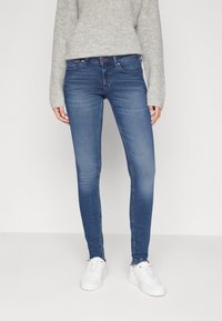 Jeans ajustados de mezclilla azul medio con un ligero desgaste, cintura media, cinco bolsillos y pasadores para cinturón, combinados con un suéter de punto gris y zapatillas blancas.