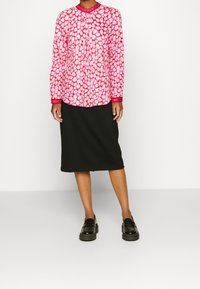 Camicia a maniche lunghe rosa con motivo rosso e bianco, abbinata a una gonna a tubino nera e mocassini neri. Tessuto dalla consistenza liscia.