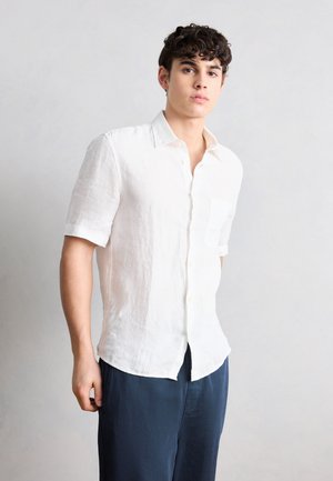 Ung mand med krøllet hår, iført en hvid kortærmet skjorte med knapper og mørkeblå bukser, stående mod en ensartet lys baggrund.
