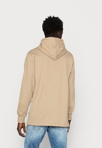 Beige hoodie med dragsko i huvan, långa ärmar och avslappnad passform. Den har en rak fåll och mjuk bomullstextur.