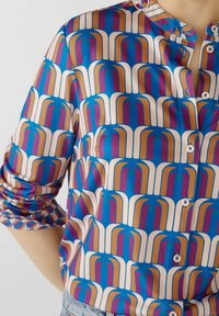 Blusa azzurra con bottoni, caratterizzata da un motivo geometrico ripetuto in bianco, arancione e rosa. Maniche corte con colletto e dettagli con bottoni.