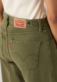 Grønne Levi's jeans med synlig baglomme, læder mærke og rød Levi's mærkat båret af en person i hvid ribbet top.