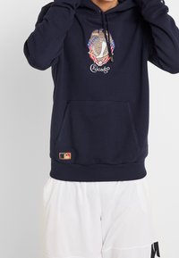 Mörkblå hoodie med en framficka i kangaroo-stil, prydd med ett broderat Chicago-emblem och MLB-logotyp vid fållen. Mjuk, texturerad tyg.