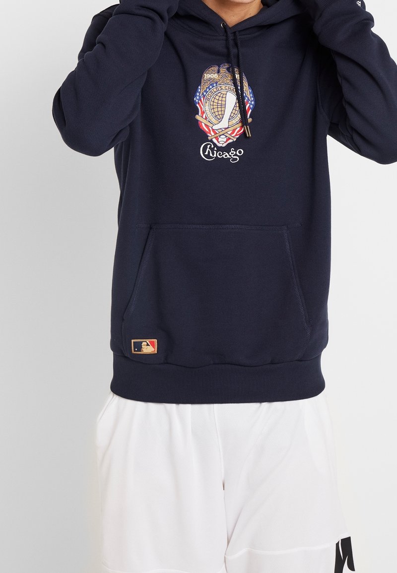 Mörkblå hoodie med en framficka i kangaroo-stil, prydd med ett broderat Chicago-emblem och MLB-logotyp vid fållen. Mjuk, texturerad tyg.