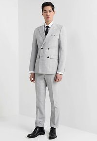 Hellgrauer doppelreihiger Anzug aus strukturiertem Stoff, mit schwarzen Knöpfen, weißem Hemd, schwarzer Krawatte und polierten schwarzen Lederschuhen.