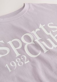 Lavender cotton t-shirt with a crew neckline, featuring large white text that reads "Sports Club 1982" prominently on the front.