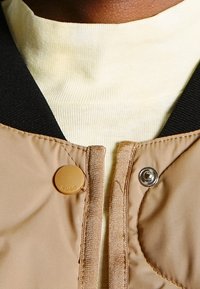 Nærbillede af beige jakke krave med sort ribbet kant, knap mærket "HUGO" og en cremefarvet ribbet turtleneck nedenunder.