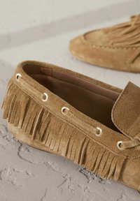 Mocassino in suede marrone chiaro con dettagli in frangia, occhielli dorati decorativi e suola piatta. Progettato per un uso facile e per il massimo comfort.