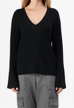 Femme portant un pull noir côtelé à col en V avec manches longues et un pantalon cargo gris avec de grandes poches.
