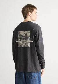 Tricou gri cu mânecă lungă, având un imprimeu pătrat de camuflaj pe spate. Textul include "Barbour International England." Croială lejeră.