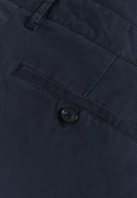 Shorts en tissu bleu marine avec une poche avant et une fermeture à bouton noir avec un logo. Texture lisse et détails de couture soignés.