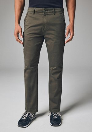 Olive green chinos with a straight fit, featuring front and back pockets. Paired with black sneakers and gray laces. Smooth fabric texture.