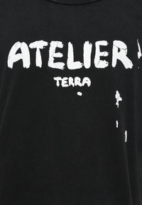 Črna bombažna majica s širokim, obrabljenim belim tiskom, ki pravi "ATELIER TERRA." Tkanina izgleda mehka z rahlo teksturo.