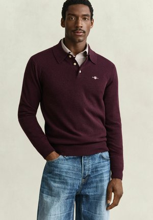 Mann trägt einen burgunderfarbenen langärmeligen Polopullover über einem hellgrauen Hemd mit blauer Jeans, steht mit einer Hand in der Tasche vor einfarbigem Hintergrund.