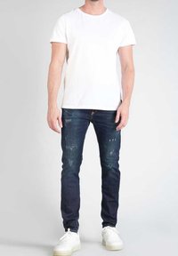 Camiseta blanca de manga corta, jeans oscuros con detalles desgastados y zapatillas blancas. Diseño simple, ajustado y con estilo casual.