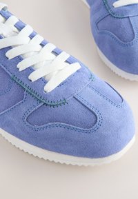 Sneakers in camoscio azzurro chiaro con lacci bianchi, decorati con cuciture verdi e suola in gomma bianca testurizzata con motivo a zigzag.