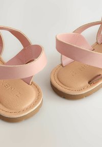 Sandali in pelle rosa con cinturini incrociati e una soletta texturizzata color tan. Accenti includono una suola beige e dettagli metallici sottili.